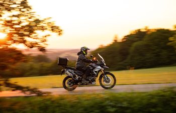 A person wearing a helmet and jacket rides a black motorcycle on a rural road at sunset, with blurred greenery in the background.