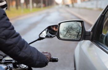 A motorcyclist wearing gloves rides close to a car, both reflected in the car's side mirror on a city street.