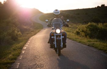 A person wearing a helmet rides a motorcycle down a rural road at sunset, with greenery on both sides and the sun low on the horizon.