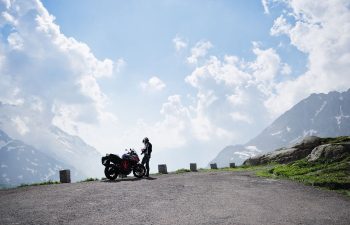 A person wearing motorcycle gear stands next to a parked motorcycle on a mountain road with cloudy sky and snow-capped peaks in the background.