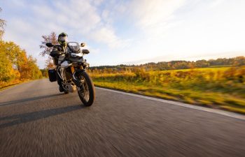 A person rides a motorcycle down a rural road with trees and fields on both sides under a clear sky.