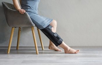 A person in a gray dress sits on a chair with a black knee brace on their right leg, foot resting on the floor.