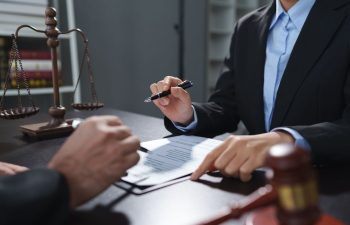 Two people in business attire discuss a document at a desk with a gavel and scales of justice, suggesting a legal or professional setting.