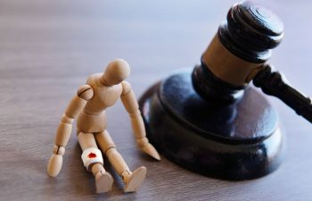 Wooden mannequin with an injured knee sits on the floor beside a judge’s gavel on a wooden surface.