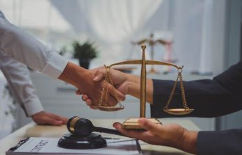 Two people shake hands across a desk with a gavel, a clipboard, and a balanced scale of justice in the foreground.