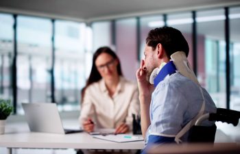 A man wearing a neck brace sits in a wheelchair, holding his face, while a woman with a laptop and notepad sits across the desk in an office setting.