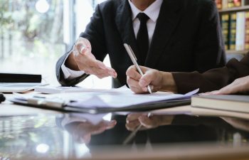 Two people in business attire sit at a desk with documents; one gestures while the other writes on a paper, suggesting a meeting or contract discussion.