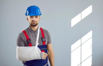 Builder gets injured at work. Portrait of man in workwear uniform, hard hat and sling immobilizer on broken arm standing on copy space wall background. Accident and injury at construction site concept